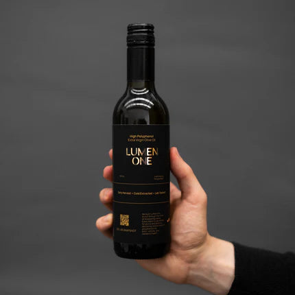 A hand holding a dark glass bottle of Lumen One high polyphenol olive oil against a dark gray background.